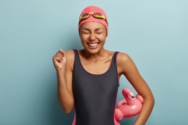 La nadadora feliz triunfante mantiene el puño cerrado, celebra la competencia ganadora, mantiene los ojos cerrados, sostiene el flamigo inflado, usa traje de baño