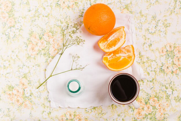 Naranja entera y rebanada; taza de café; Botella de leche y gypsophila sobre fondo floral.
