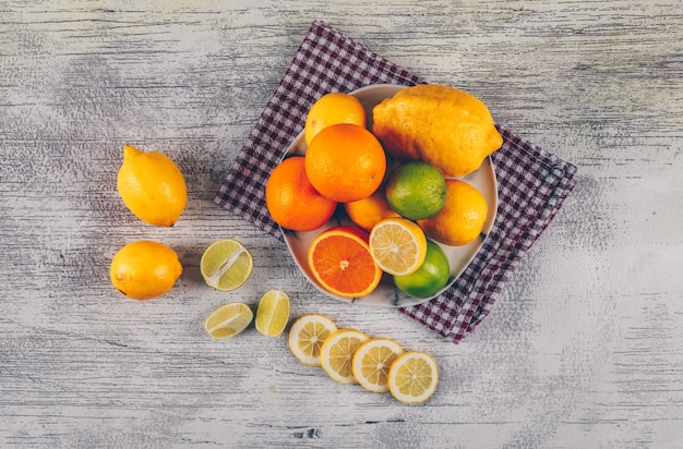 Naranja con limones verdes y amarillos con rodajas en un plato sobre tela y fondo de madera gris, vista superior.