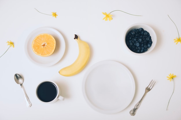 Naranja a la mitad plátano; cuenco de arándanos; Taza de café y un plato vacío sobre fondo blanco