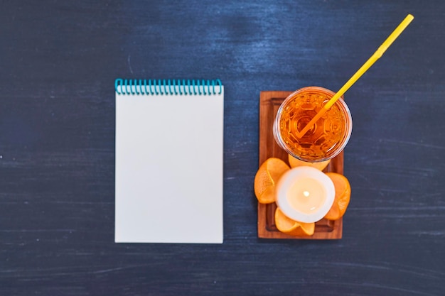 Naranja y un vaso de jugo con tubo amarillo en bandeja de madera con cuaderno blanco. Foto de alta calidad