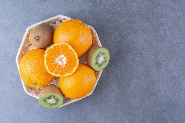 Naranjas y kiwis en canasta sobre mesa de mármol.