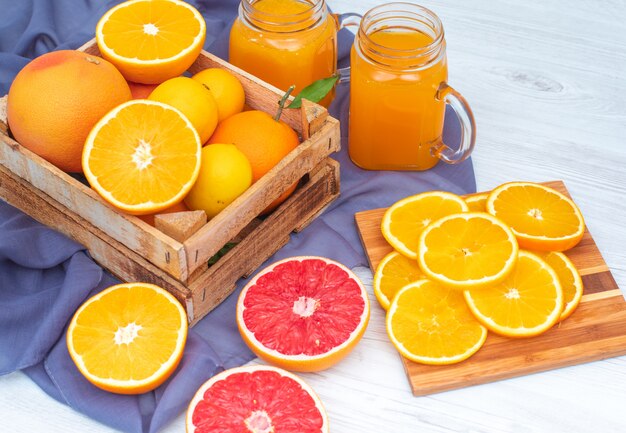 Naranjas y limones en la caja de madera frente a vasos de jugo de naranja sobre tela violeta
