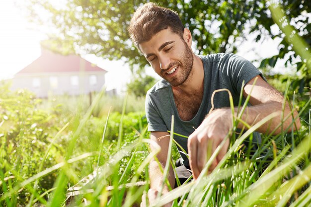 Naturaleza y Medio Ambiente. Joven jardinero barbudo de piel oscura pasar tiempo en el jardín cerca de la casa de campo. Hombre cortando hojas y disfrutando del clima caluroso de verano en la sombra de los árboles