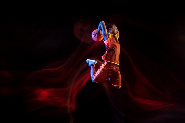 Naturaleza misteriosa. Joven jugador de baloncesto afroamericano del equipo rojo en acción y luces de neón sobre fondo oscuro de estudio. Concepto de deporte, movimiento, energía y estilo de vida dinámico y saludable.