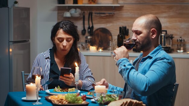 Navegando en teléfonos durante la cena Pareja sosteniendo teléfonos inteligentes en la cocina sentado en la mesa navegando, buscando, usando teléfonos inteligentes, Internet, celebrando su aniversario en el comedor.
