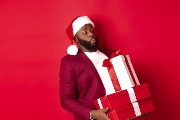 Navidad, año nuevo y concepto de compras. Gracioso hombre afroamericano con sombrero de santa llevar regalos de Navidad pesados, sosteniendo regalos, de pie sobre fondo rojo.