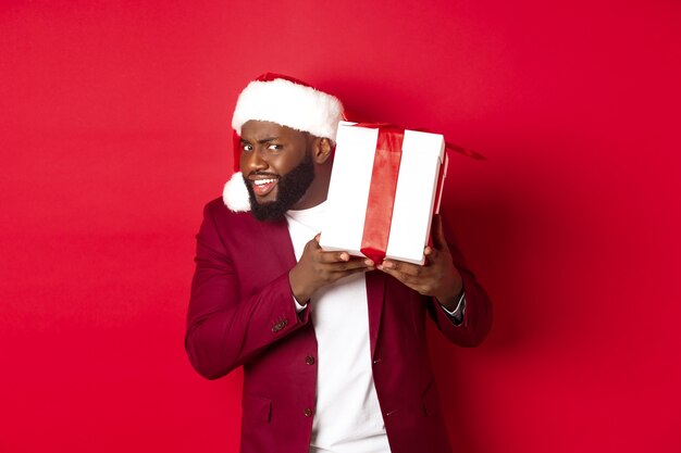 Navidad. Curioso hombre negro con sombrero de santa agitando regalo de año nuevo