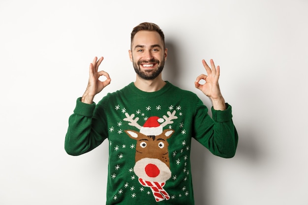 Navidad, fiestas y celebraciones. Hombre sonriente satisfecho con suéter verde que muestra signos de OK y asintiendo con la cabeza en señal de aprobación, recomendando el producto, de pie sobre un fondo blanco.