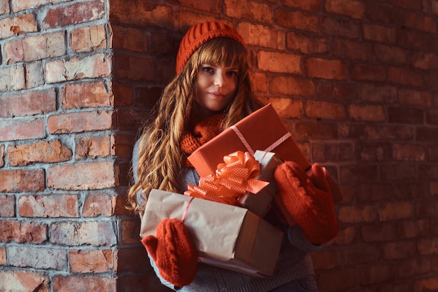 Navidad, San Valentín, Año Nuevo. Retrato de una pelirroja solitaria con un suéter cálido y un sombrero sosteniendo cajas de regalo mientras se apoya en una pared de ladrillos.