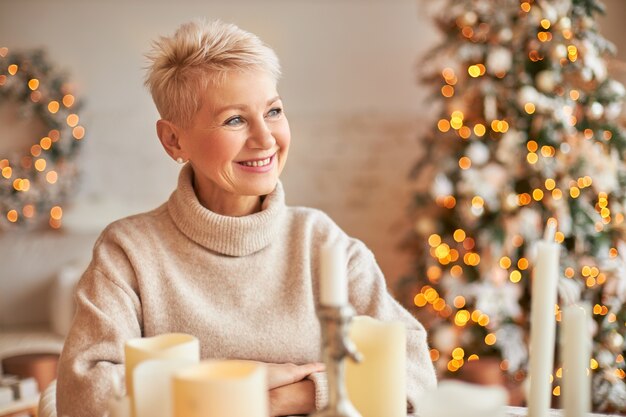 Navidad, vacaciones, decoración, fiesta y concepto de ambiente festivo. Atractiva mujer de mediana edad alegre con pelo corto disfrutando del humor navideño, sentado alrededor de velas de cera, adornos y luces