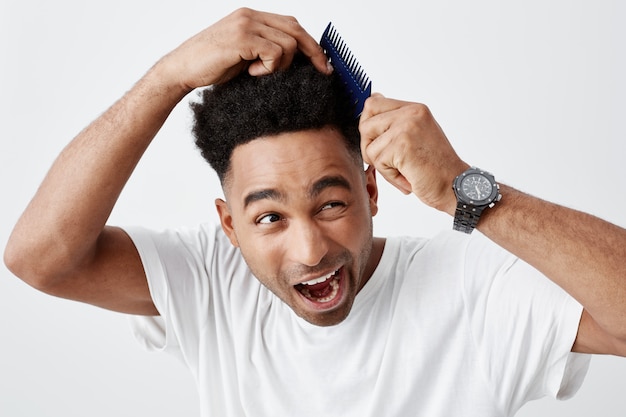 Necesita nuevo corte de pelo. Retrato de un chico africano maduro, divertido, hermoso, de piel bronceada, con cabello rizado en una camiseta blanca informal que tiene problemas para peinarse, luciendo asustado con expresión dolorosa