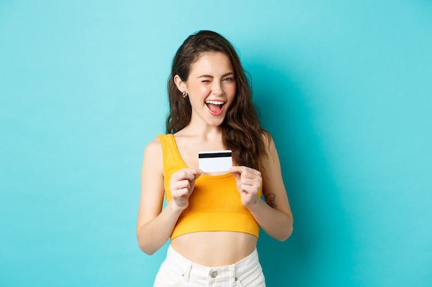 Necesitas esto. Sonriente mujer atractiva insinuando, guiñando un ojo y mostrando una tarjeta de crédito de plástico, recomendando el pago sin contacto, de pie sobre fondo azul.