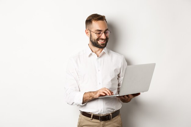 Negocio. Apuesto hombre de negocios trabajando en equipo portátil, respondiendo mensajes y sonriendo, de pie