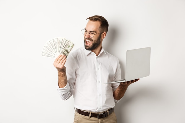 Negocios y comercio electrónico. Hombre de negocios exitoso usando laptop para trabajar y sosteniendo dinero, de pie