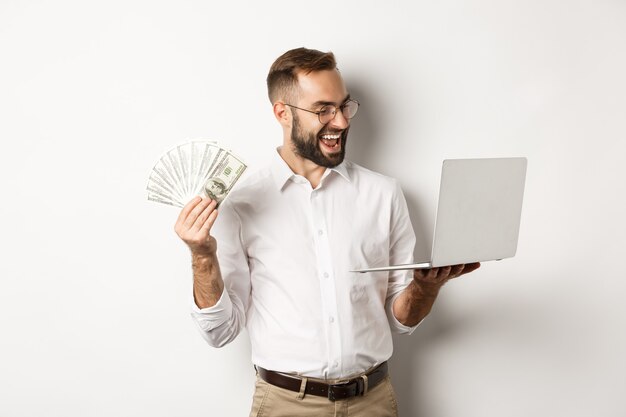 Negocios y comercio electrónico. Hombre de negocios satisfecho haciendo trabajo en la computadora portátil y sosteniendo dinero, sonriendo feliz, de pie