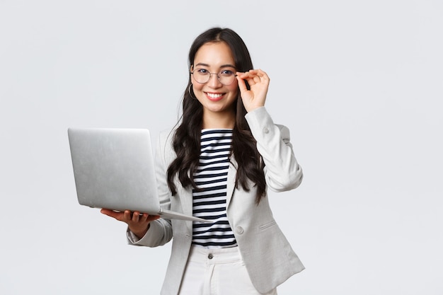 Negocios, finanzas y empleo, concepto de mujeres emprendedoras exitosas. Confianza empresaria asiática sonriente, oficinista con traje blanco y gafas usando una computadora portátil, ayuda a los clientes