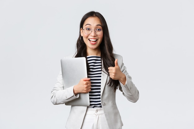 Foto gratuita negocios, finanzas y empleo, concepto de mujeres emprendedoras exitosas. joven empresaria segura de anteojos, mostrando gesto de pulgar hacia arriba, sostenga la computadora portátil, garantice la mejor calidad de servicio