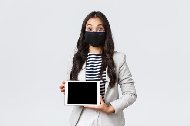 Negocios, finanzas y empleo, covid-19 previniendo el virus y el concepto de distanciamiento social. Emocionada empresaria asiática en mascarilla y gafas abren los ojos sorprendidos, mostrando la pantalla de la tableta digital