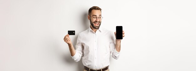 Negocios y pago en línea hombre guapo sonriente que muestra la pantalla del móvil y la tarjeta de crédito de pie