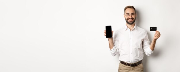 Negocios y pago en línea hombre guapo sonriente que muestra la pantalla del móvil y la tarjeta de crédito de pie