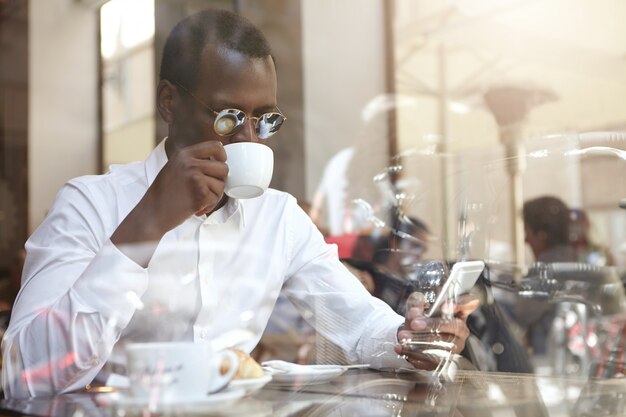 Negocios, tecnología moderna, comunicación y concepto de personas. Hombre de negocios afroamericano elegante confiado en gafas de sol redondas bebiendo cappccino en la cafetería, sentado junto a la ventana con teléfono celular