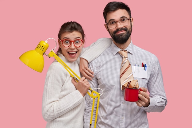 Los nerds sonrientes y alegres de la mujer joven y del hombre se paran cerca uno del otro, colaboran para preparar y terminar la tarea común, sostienen la lámpara de mesa y la taza con bebida