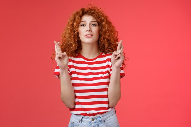 Nerviosa joven tonta tímida linda pelirroja pelirroja anticipando resultados esperanzadores creer rezando entrecerrar los ojos intensa preocupación para ganar los dedos cruzados buena suerte deseo hecho realidad fondo rojo.