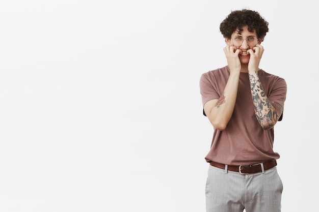 Foto gratuita nervioso joven y elegante chico de pelo rizado que tiene el mal hábito de morderse las uñas y fruncir el ceño, comenzando a sentir pánico mientras espera una llamada importante, preocupado de pie sobre una pared gris