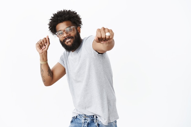 Únete a mí en la competencia de baile. Retrato de feliz chico afroamericano apuesto enegized con gafas con anillo y nariz perforada sonriendo tirando del puño hacia la cámara mientras baila, dándose la mano