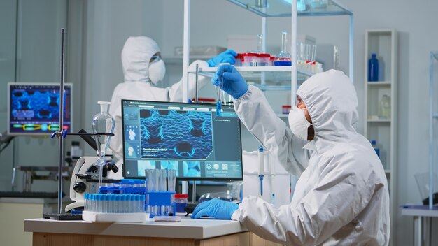 Neurólogo con traje de ppe trabajando en el desarrollo de vacunas en un laboratorio equipado escribiendo en la pc. Equipo que examina la evolución del virus utilizando alta tecnología para la investigación en el desarrollo de tratamientos contra covid19