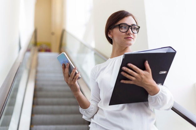 Ángulo bajo, de, mujer de negocios, con, carpeta, y, smartphone, en, escalera mecánica