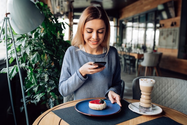 Nicegirl alegre que toma la foto del pastel sabroso en el teléfono móvil, pasando tiempo en el café