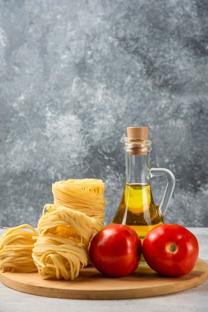 Nidos de pasta cruda, botella de aceite de oliva y tomates en placa de madera.