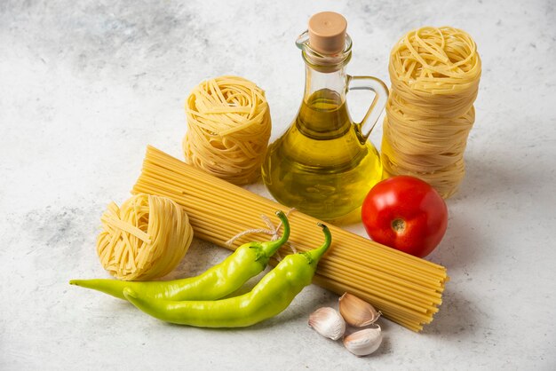 Nidos de pasta cruda, espaguetis, botella de aceite de oliva y verduras sobre superficie blanca.