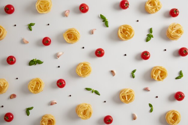 Nidos de pasta cruda hechos de harina de trigo duro, tomates maduros, ajo, hojas de albahaca y granos de pimienta para preparar pasta. Comida italiana, concepto de cocina. Alimentación nutritiva. Fideos sobre fondo blanco