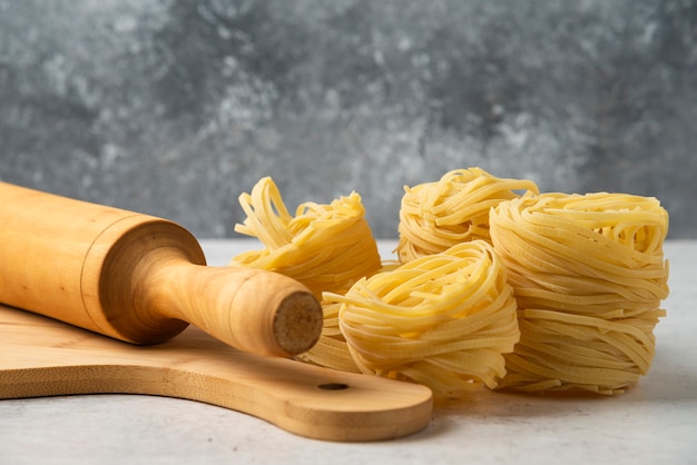 Foto gratuita nidos de pasta cruda, tablero de madera y rodillo sobre mesa blanca.