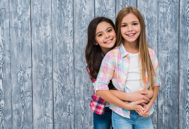 Niña abrazando a su mejor amiga por detrás, de pie contra la pared de madera gris