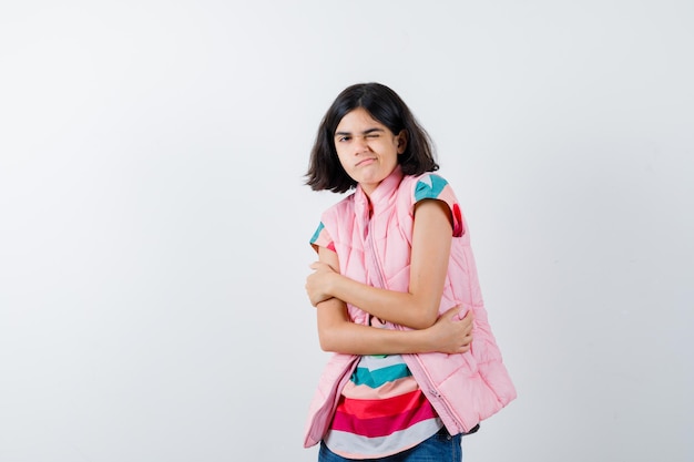 Niña abrazándose a sí misma en camiseta, chaleco acolchado, jeans y mirando perplejo, vista frontal.