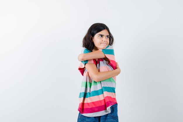 Niña abrazándose a sí misma, mirando a otro lado con una camiseta a rayas de colores y luciendo seria. vista frontal.