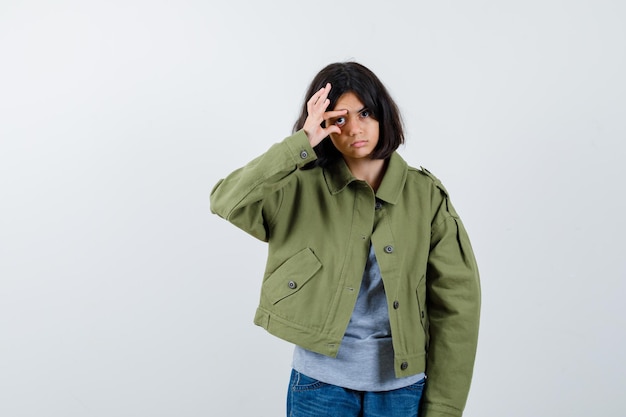 Niña abriendo el ojo con los dedos en el abrigo, camiseta, jeans y luciendo linda vista frontal.