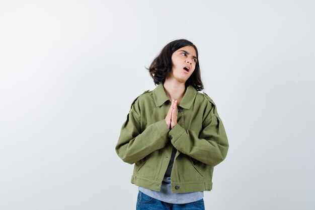 Niña con abrigo, camiseta, jeans presionando las manos juntas para orar y mirando esperanzada, vista frontal.