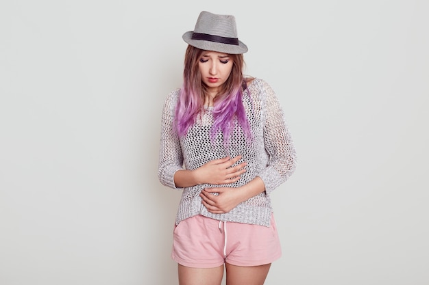 Foto gratuita niña adulta joven con cabello lila con sombrero que tiene problemas con el estómago, que sufre de dolor menstrual, manteniendo las manos en el vientre, aislado sobre una pared blanca.