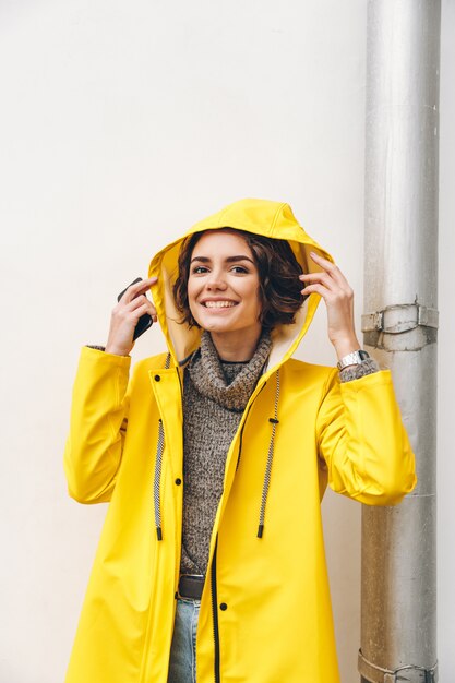 Niña adulta sonriente en capa amarilla poniendo capucha en la cabeza posando en cámara con sincera sonrisa ser feliz