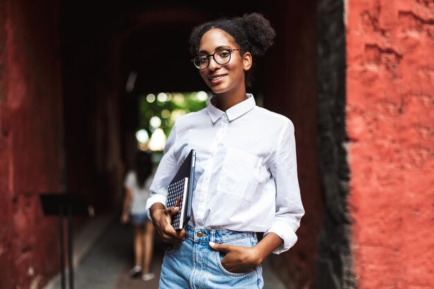 Niña africana muy sonriente con anteojos y camisa sosteniendo una computadora portátil y un bloc de notas en las manos mientras mira soñadoramente en la cámara al aire libre