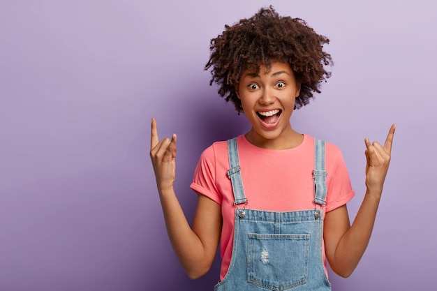 La niña afroamericana despreocupada y alegre grita sí, muestra un gesto de rock n roll con ambas manos, mantiene la boca abierta, se viste con ropa elegante, posa sobre un fondo púrpura