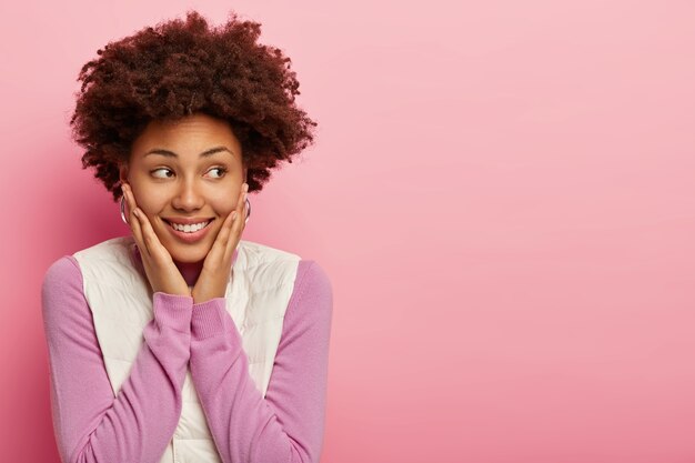 Una niña afroamericana feliz de aspecto amistoso mira a un lado con una sonrisa y sorpresa, toca ambas mejillas con las palmas, usa un jersey púrpura y un chaleco blanco, tiene una expresión cálida y tierna, aislada en una pared rosa