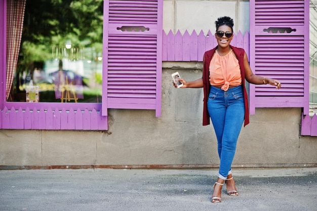 Niña afroamericana con gafas de sol posadas contra ventanas moradas con teléfono celular a mano