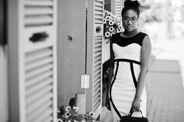 Niña afroamericana con gafas, vestido blanco y negro y bolso posado contra ventanas moradas