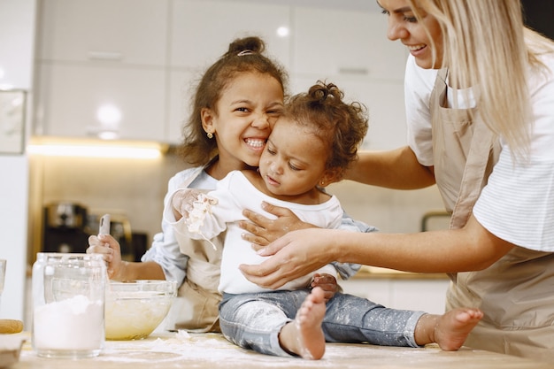 Foto gratuita niña afroamericana mezclando la masa en un recipiente de vidrio, preparando un pastel. su hermana pequeña sentada en una mesa. su madre les enseña.